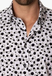 Camisa blanca con un patrón de lunares en círculos negros y verdes. La tela es suave, con un cuello abotonado y un detalle del logo en el pecho.
