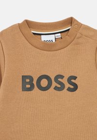Smeđi pamučni majica s okruglim izrezom, s velikim crnim "BOSS" logom sprijeda i detaljem s dugmetom blizu ramena.