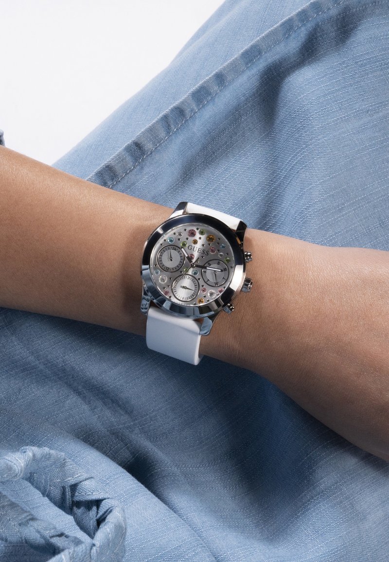 Poignet portant une montre Guess en argent avec bracelet blanc, cadran à multiples compteurs et accents de pierres précieuses colorées sur un fond en denim bleu clair.