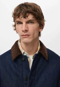 Modra denim jakna z rjavim kordurovim ovratnikom, gumbi spredaj in plastno karirasto srajco spodaj. Osredotočenost na teksturo tkanine in kontrast barv.