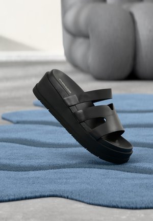 Sandalo ciabatta piattaforma nero con tre cinturini, posato su un tappeto blu con trama ondulata, all'interno vicino a un divano rotondo sfocato.