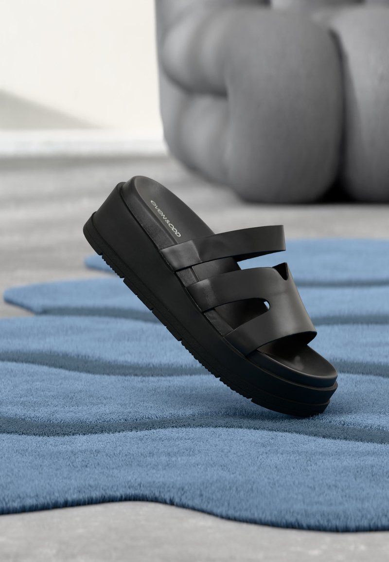 Μαύρο πέδιλο πλατφόρμας με τρεις λουριά, ακουμπισμένο σε μπλε χαλί με κυματιστή υφή, σε εσωτερικό χώρο κοντά σε θολό, στρογγυλεμένο καναπέ.