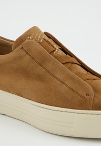 Brauner Wildledersneaker mit runder Zehenpartie, ausgestattet mit einer cremefarbenen Gummisohle, geflochtenen Schnürsenkeln und einem strukturierten Seitenpaneel. Goldfarbene Akzente am Obermaterial.