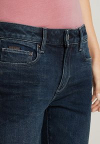 Nahaufnahme von dunkelblauen Denim-Jeans an der Taille und dem oberen Oberschenkel, getragen mit einem pinken Oberteil. Die Knöpfe, Gürtelschlaufen und Fronttaschen sind gut sichtbar.
