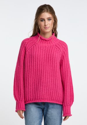 Junge Frau mit langen Haaren trägt einen knallpinkfarbenen grob gestrickten Pullover mit Ballonärmeln und blaue Jeans, steht vor einem schlichten Hintergrund.