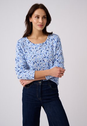 Donna che indossa una maglietta a maniche lunghe con motivo blu e bianco e jeans blu scuro, in piedi con le braccia incrociate davanti a uno sfondo semplice.