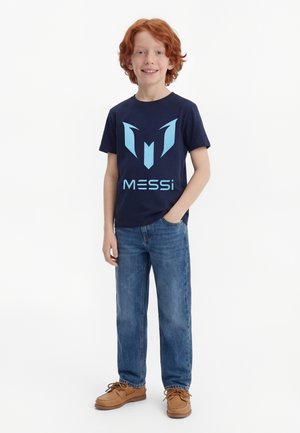 Rdečelasi deček se smeji, nosi temnomodro majico z grafiko Messija, modre kavbojke in rjave čipkaste čevlje ter stoji z eno roko v žepu.