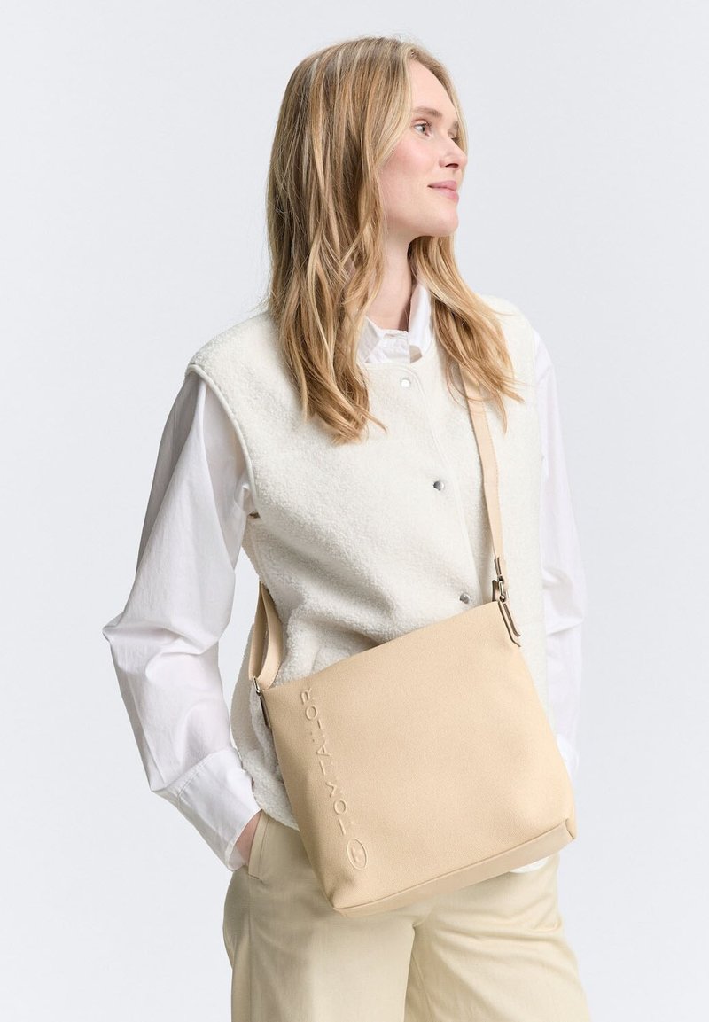 Beżowa torebka na ramię o gładkiej fakturze, z wytłoczonym logo marki. Kwadratowy kształt, regulowany pasek i minimalistyczny design.