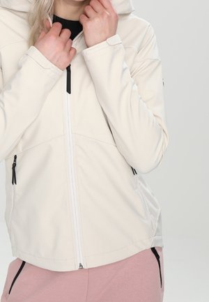 Personne portant une veste zippée beige clair avec capuche et tirettes de fermeture éclair noires sur un t-shirt noir et un pantalon rose. Mains près du col.