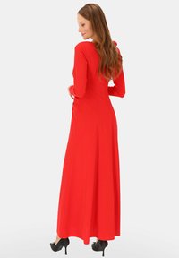 Czerwona sukienka z długim rękawem wykonana z gładkiej tkaniny, dopasowana w górnej części, z rozkloszowanym dołem i detalami w postaci szwów. Noszona z czarnymi butami na wysokim obcasie.
