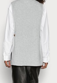 Gilet tricoté gris clair avec une finition texturée. Présente un col arrondi, des côtés ouverts et des boutons en bois au dos.