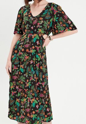 Femme portant une robe midi noire à décolleté en V avec un imprimé floral multicolore vif et manches courtes, main sur la hanche, portant des bracelets.