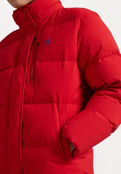 Crvena puffer jakna s visokim kragnom, horizontalnim šavovima i zip zatvaračem na prsnom džepu s plavim logom. Glatka tekstura.