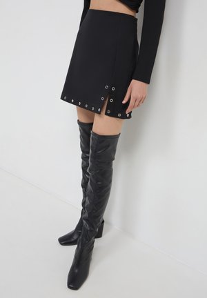 Femme portant une mini-jupe noire avec des œillets en métal et des bottes en cuir noir jusqu'à la cuisse, se tenant devant un mur blanc.