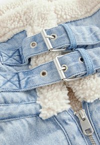 Giacca di jeans in tessuto blu chiaro, con un morbido rivestimento in pile crema e accessori in argento, tra cui fibbie e una cerniera.