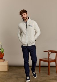 Grijze zip-up sweatshirt met hoge kraag, featuring geborduurde logo-details. Gecombineerd met donkerblauwe broek en marineblauwe sneakers.