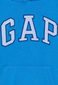 Sininen puuvillapusero, jossa on suuri valkoinen "GAP"-teksti tummansinisellä ääriviivalla. Sisältää etutaskun ja pehmeää, teksturoitua kangasta.