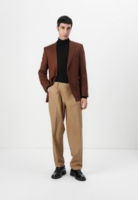 Rjav tailored blazer, črna polovična majica, bež nabirane hlače in črne čevlje. Gladke teksture in čiste linije. Enostavna obleka na enobarvni podlagi.