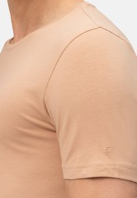 Lichtbruine getailleerde T-shirt met korte mouwen, ronde halslijn en subtiele logodetail op de mouw. Soepel, zacht stofoppervlak.