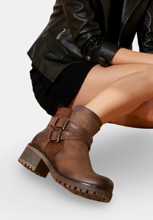 Bottines en cuir marron avec des doubles sangles à boucle, portées avec une jupe noire et une veste en cuir noire, mannequin assis les jambes croisées.