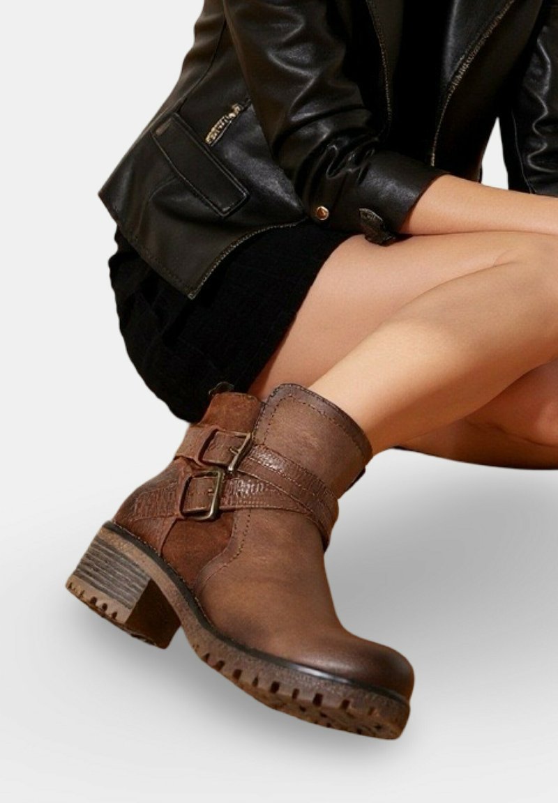 Bottines en cuir marron avec des doubles sangles à boucle, portées avec une jupe noire et une veste en cuir noire, mannequin assis les jambes croisées.