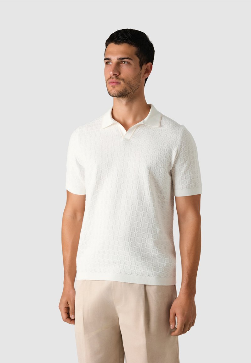 Homme portant un polo à manches courtes texturé blanc et un pantalon beige, debout devant un fond gris clair uni.