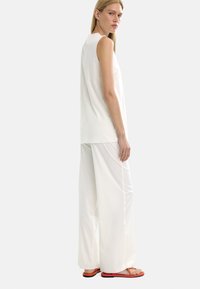 Biała bezrękawnikowa bluzka w połączeniu z luźnymi białymi spodniami. Gładka tekstura i swobodny krój. Pomarańczowe płaskie sandały dopełniają stylizację.