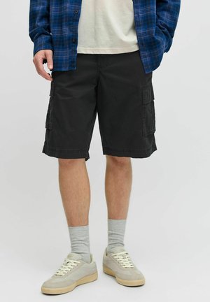 Mann trägt schwarze Cargoshorts, graue Sportsocken, beige Sneakers, weißes T-Shirt und blau kariertes Hemd mit der Hand in der Tasche.