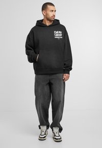 Svart hettegenser med frontlomme, med hvit tekst "Ring min advokat" og et telefonnummer. Brukt med løse grå jeans og sneakers.