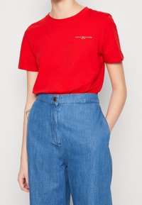 Rød bomulds T-shirt med rund halsudskæring og korte ærmer, der har et lille hvidt logo på venstre bryst. Sammen med højtaljede blå denimbukser.