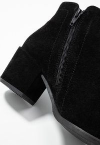 Botín negro de ante hasta el tobillo con un tacón cuadrado y grueso y cremallera lateral. Textura suave y diseño moderno, ideal para un estilo versátil.