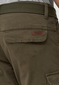 Olive green pants with a woven belt, featuring a leather patch labeled "INDICODE" and a back pocket with stitching details.