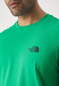 Vihreä puuvillapaita, jossa on pyöreä pääntie ja lyhyet hihat. Musta logo "The North Face" vasemmassa rintakotelossa. Suhdeena sileä tekstuuri.