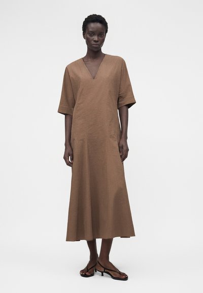 Γυναίκα με καφέ φόρεμα με V-λαιμόκοψη, κοντά μανίκια, μήκος μέχρι τους αστραγάλους και μαύρα σανδάλια με λεπτά λουράκια και χαμηλό τακούνι, στέκεται μπροστά σε απλό φόντο.