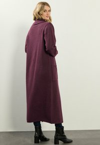 Lång, djup lila cardigan tillverkad av mjukt stickat tyg med sjalkrage och två sidofickor. Bärs över mörka jeans och svarta ankelstövlar.