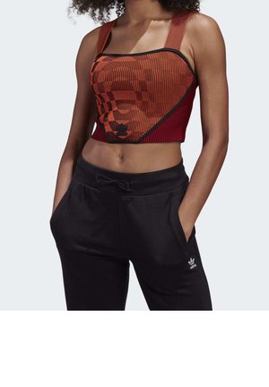 Vrouw draagt een rood-zwart patroon crop top en zwarte Adidas trainingsbroek met handen in de zakken, tegen een effen achtergrond.