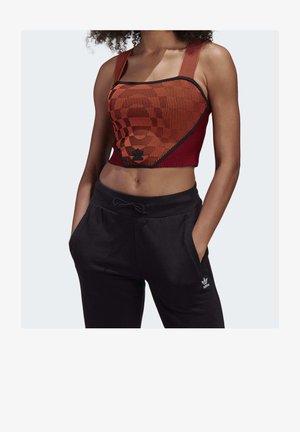 Femme portant un crop top à motifs rouge et noir et un pantalon de survêtement Adidas noir, les mains dans les poches, sur un fond uni.