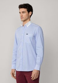 Camisa de botones azul claro con rayas verticales blancas y naranjas, cuello blanco, bolsillo en el lado izquierdo y mangas largas. Material de algodón.