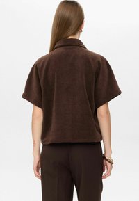 Woman with long hair wearing a loose dark brown short-sleeve top and matching pants, standing facing away, showing the back of the outfit.