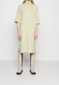 Knitklänning i blekgul med vågiga vita mönster, har högringad hals och korta ärmar; sidospalt synlig. Buren med beigefärgade knähöga stövlar.