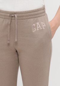 Pantaloni de trening maro deschis cu talie elastică, șnur, buzunare laterale și logo-ul "GAP" brodat într-o culoare contrastantă.