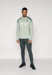 Svetlosivý športový top s dlhým rukávom a čiernymi akcentmi, s logom a textom „Bingx“. Doplnok k tmavozeleným úzkym nohaviciam a čiernym topánkam.