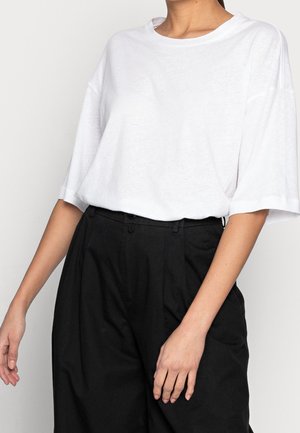 Femme portant un t-shirt blanc à manches courtes ample, rentré dans un pantalon noir taille haute à jambes larges, montrée des épaules aux hanches.