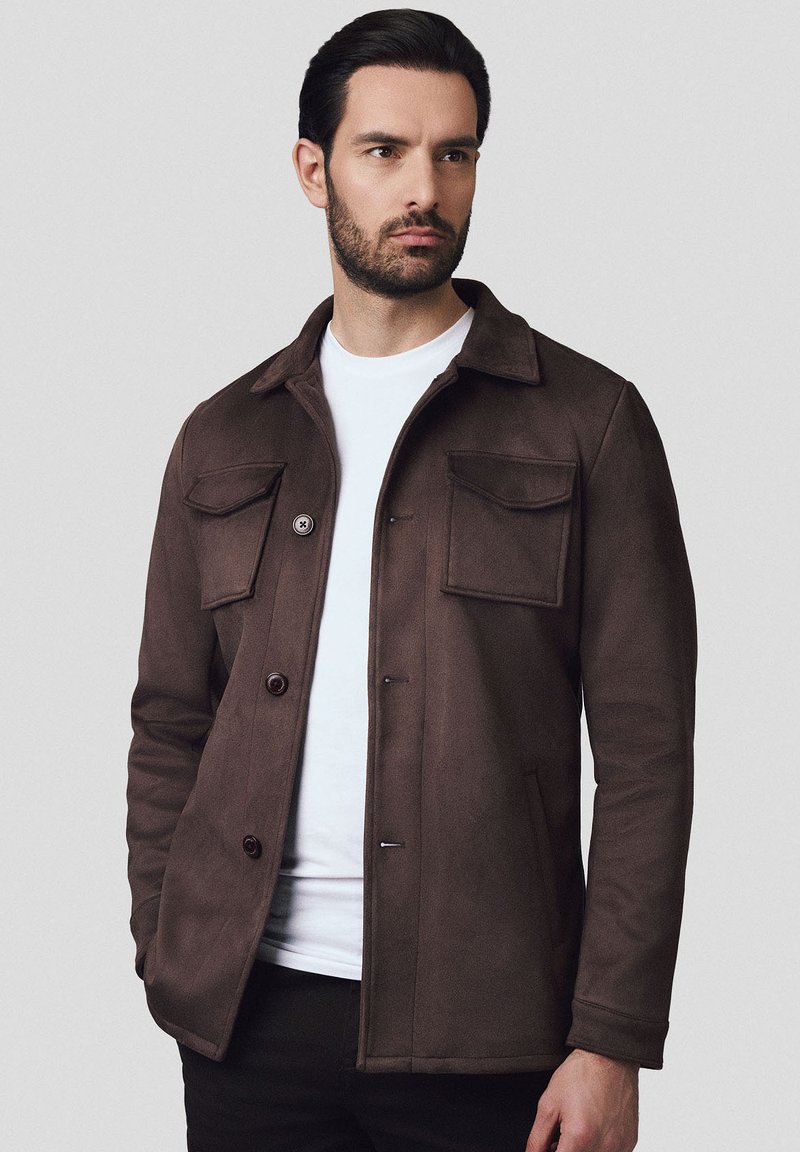 Brązowa kurtka z gładkiego materiału, z kołnierzem, dwoma przednimi kieszeniami, zapięciem na guziki oraz dopasowanym krojem na białej koszuli.