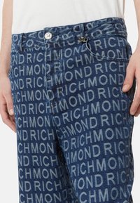Pantaloni in denim con una stampa "RICHMOND" su tutta la superficie in diverse dimensioni. Il tessuto è di colore blu scuro con una texture liscia e una leggera lucentezza.