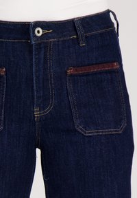 Donkerblauwe denim jeans met een hoge taille, zichtbare gele steken en een bruin accent op de achterzak. Knopsluiting bij de taille.