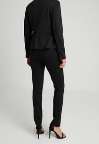 Blazer noir sur mesure avec ourlet évasé et pantalon noir ajusté. Porté avec des chaussures à talons ouverts. Tissu lisse et structuré.