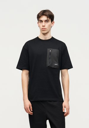 Mladi moški v črni majici s kratkimi rokavi z velikim žepom z zadrgo na levi strani prsnega koša, ki stoji pred enostavnim svetlim ozadjem.