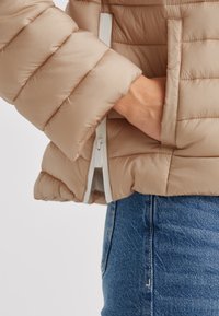 Beige Steppdaunenjacke mit gestepptem Muster und elastischen Bündchen, ausgestattet mit einer seitlichen Reißverschlusstasche und einem kontrastierenden weißen Reißverschluss. Darunter sind blaue Jeans sichtbar.