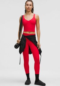 Mujer con top rojo y leggings sosteniendo una esterilla de yoga enrollada, con una chaqueta negra atada a la cintura, usando calcetines negros y zapatos tipo slip-on.